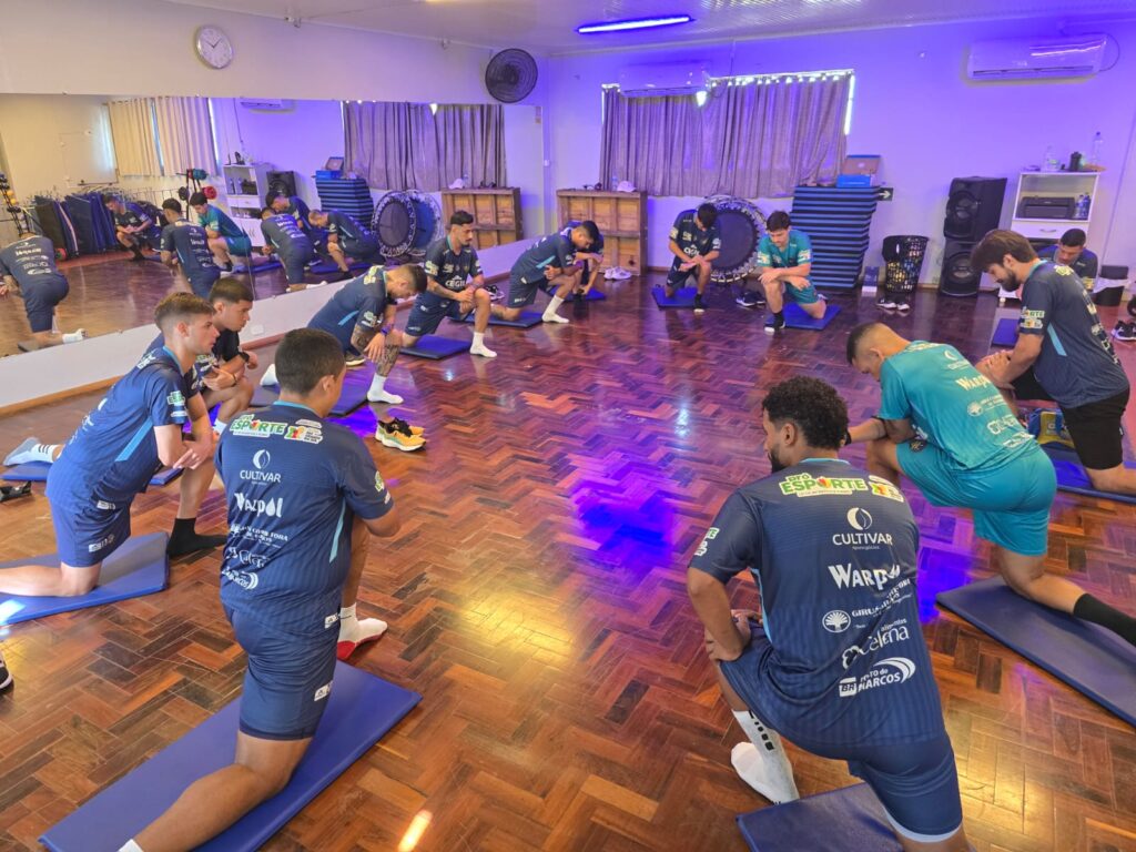 Jogadores de um time de futebol realizam alongamentos em grupo dentro de uma sala de treino com piso de madeira. Eles estão ajoelhados em colchonetes azuis, dispostos em um grande círculo, vestindo uniformes de treino azuis com diversos logotipos de patrocinadores.