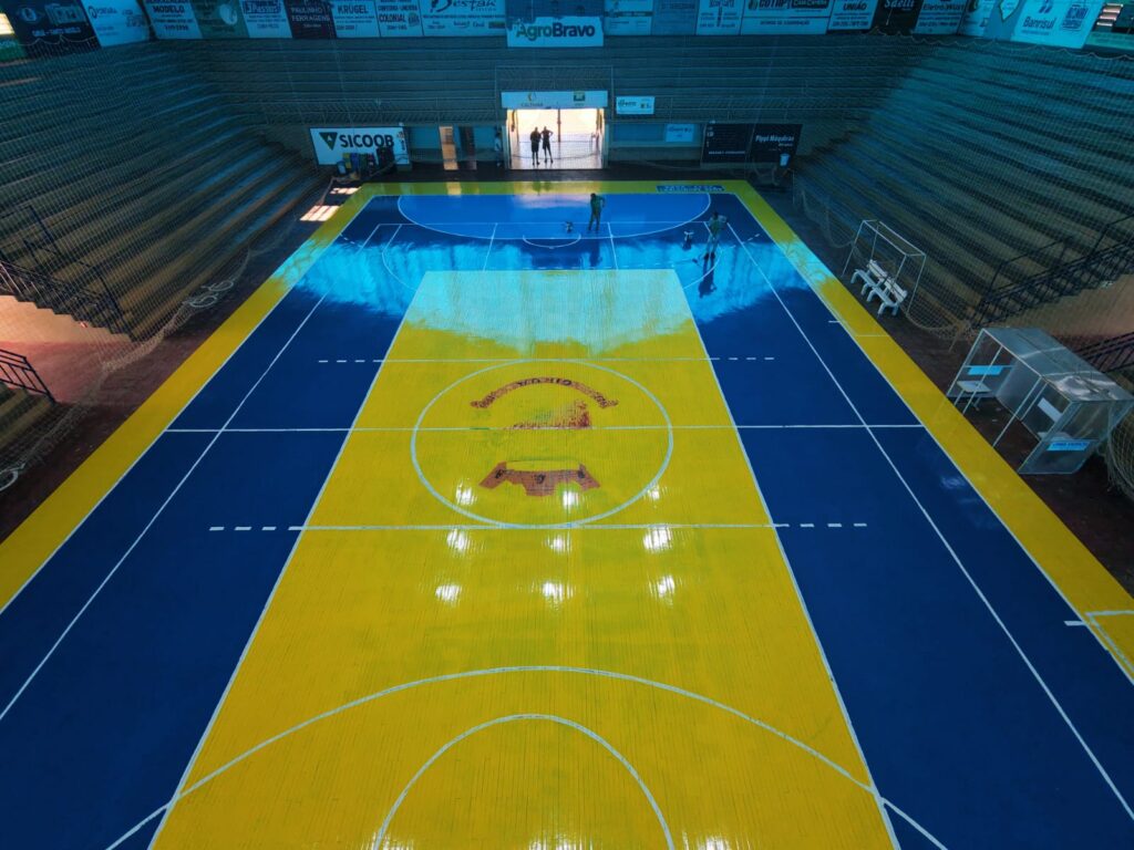 Vista aérea de uma quadra poliesportiva interna com piso de madeira sendo revitalizado. O centro da quadra é amarelo vibrante com um escudo circular, e as laterais e áreas de garrafão são azul-escuro, tudo com um acabamento de verniz altamente brilhante e reflexivo.