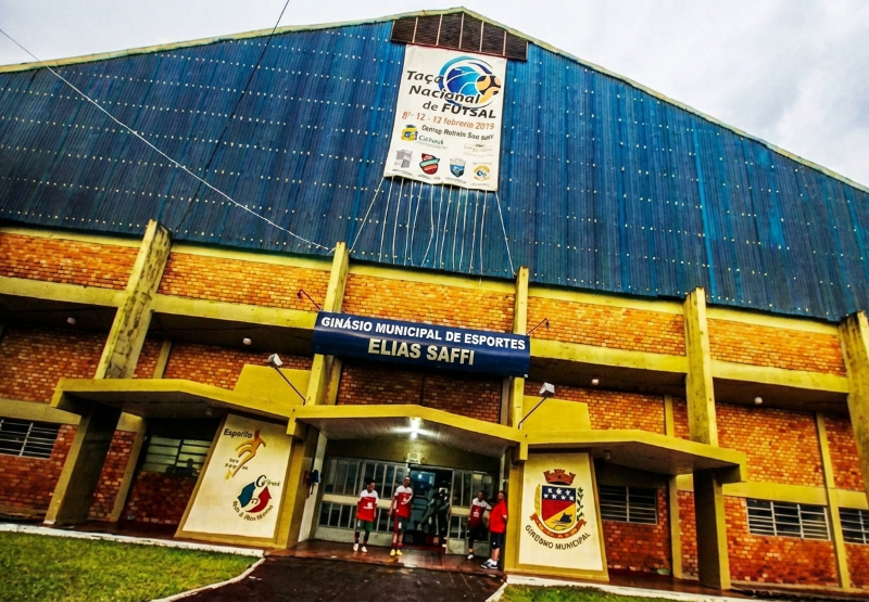 Fachada externa do Ginásio Municipal de Esportes Elias Saffi, um prédio de tijolos aparentes com uma grande estrutura azul na parte superior. Uma faixa vertical branca e azul está pendurada no centro, anunciando a 'Taça Nacional de Futsal', com datas e logotipos. O nome do ginásio está escrito acima da entrada principal, onde algumas pessoas estão reunidas.